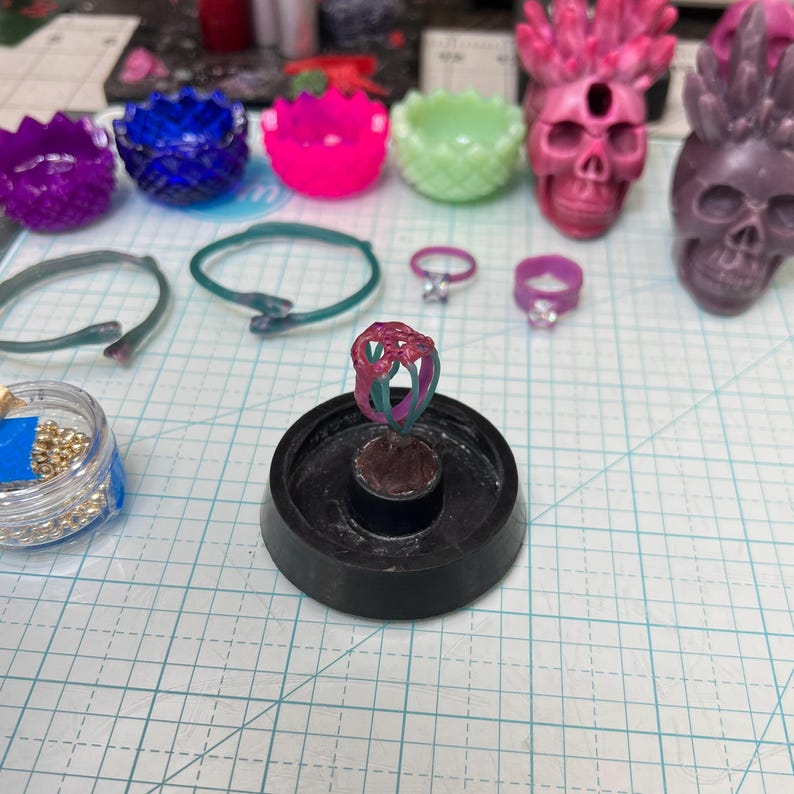 May include: A collection of jewelry and decorative items, including rings, bracelets, and skull-shaped figurines. The rings are in various colors, including pink and green. Other items include small, colorful bowls. The items are on a grid-lined surface.
