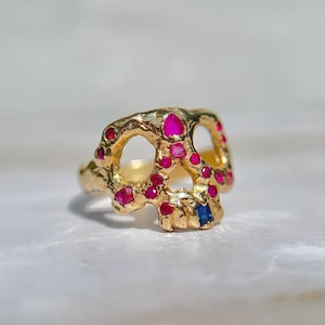 May include: A gold-colored ring in the shape of a skull, adorned with red and pink gemstones. A single blue gemstone is set in the lower jaw. The ring has a textured surface and is set against a white background.
