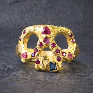 May include: A gold-colored skull ring with ruby and sapphire accents. The ring features a skull design with open eye sockets and is adorned with numerous red gemstones and a single blue gemstone.