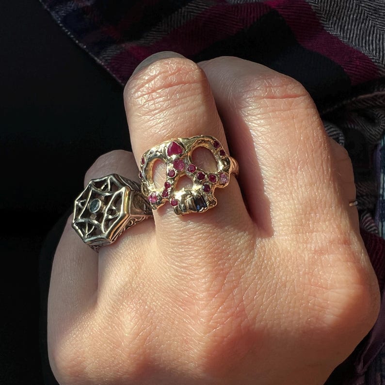 May include: Two rings are displayed. One is gold-toned and shaped like a skull, adorned with red and purple gemstones. The other ring is silver-toned with an octagonal design and a central green stone. Both rings are on a hand.