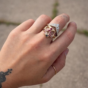 May include: A hand wearing several gold rings. One ring is a skull design with red and black gemstones. Another ring is a chevron shape with clear gemstones. A simple gold band is also visible.