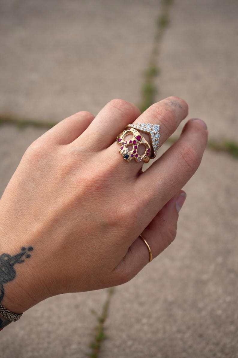 May include: A hand wearing several gold rings. One ring is a skull design with red and black gemstones. Another ring is a chevron shape with clear gemstones. A simple gold band is also visible.