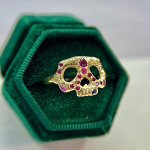 May include: A gold skull ring with ruby and sapphire accents. The ring is set with multiple red rubies and a single blue sapphire, arranged to resemble a skull. The ring is displayed in a green velvet ring box.