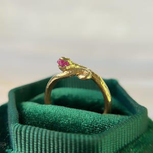 Op de afbeelding: Een goudkleurige ring met een getextureerde band en een kleine, helder roze edelsteen. De ring heeft een uniek, organisch ontwerp, mogelijk geïnspireerd door de natuur. De ring wordt gepresenteerd in een groene fluwelen doos.
