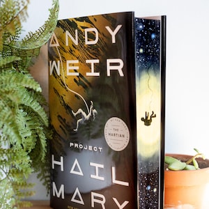 May include: A hardcover book titled "Project Hail Mary" by Andy Weir. The cover features an astronaut figure and a gold and black design. The book is a New York Times bestseller. A plant and a small pot are in the background.