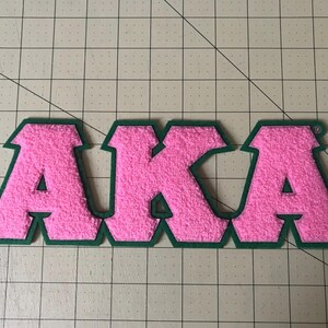 May include: A pink felt patch with the letters "AKA" in a bold, blocky font. The letters are outlined in green, creating a contrasting border. The patch is likely for apparel or accessories.