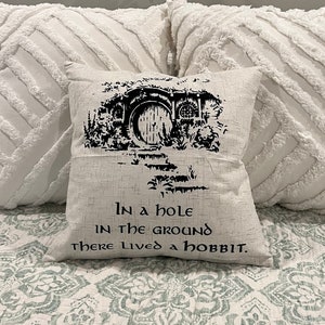 Puede incluir: Una almohada blanca con texto negro que dice "In a hole in the ground there lived a hobbit." La almohada presenta una ilustración en blanco y negro de una madriguera de hobbit con una puerta redonda.