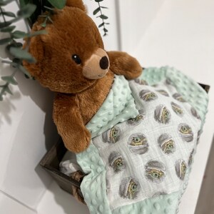 May include: A brown teddy bear is lying in a green and white baby blanket with a repeating pattern of a cartoon character. The blanket has a soft, textured feel.