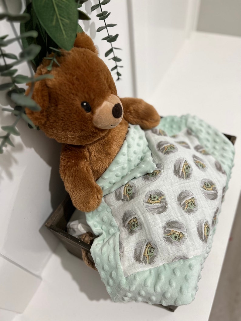 May include: A brown teddy bear is lying in a green and white baby blanket with a repeating pattern of a cartoon character. The blanket has a soft, textured feel.