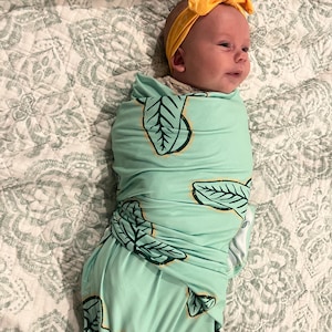 May include: A baby swaddled in a mint green swaddle with a leaf pattern. The baby is wearing a yellow headband.