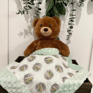 May include: A brown teddy bear is lying under a green and white baby blanket with a repeating pattern of a cartoon character. The blanket has a soft, textured border.