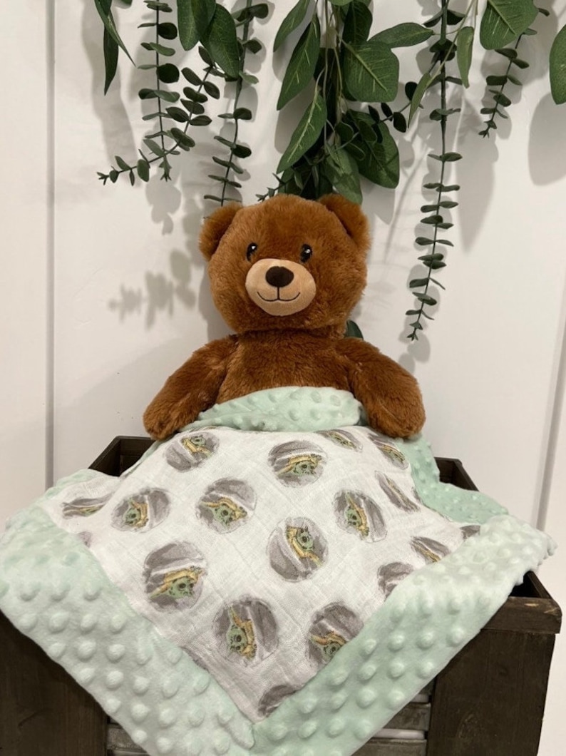May include: A brown teddy bear is lying under a green and white baby blanket with a repeating pattern of a cartoon character. The blanket has a soft, textured border.