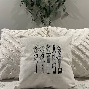 Puede incluir: Una almohada blanca con un diseño negro de cinco sables de luz con flores que crecen desde la parte superior. Los sables de luz están en fila y las flores son de diferentes tipos.