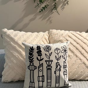 Puede incluir: Un cojín cuadrado de color claro con un diseño floral en blanco y negro. El diseño presenta flores estilizadas sobre lo que parecen ser empuñaduras de sables de luz. El cojín está sobre una cama con otros dos cojines blancos.