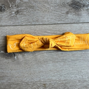 May include: A yellow headband with a white design of the inscription "One Ring to rule them all" from the Lord of the Rings.