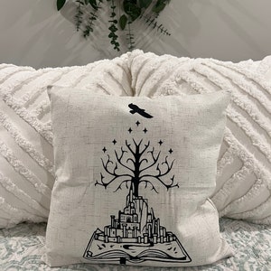 May include: A white throw pillow with a black design of a tree growing out of a book with a city on top. A black bird is flying above the tree.