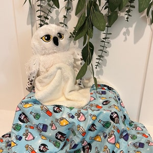 Könnte beinhalten: Ein weißes Plüsch-Eulenspielzeug mit schwarzen Augen und einem braunen Schnabel liegt auf einer blauen und weißen Decke mit einem Muster aus bunten Zaubertrankflaschen.