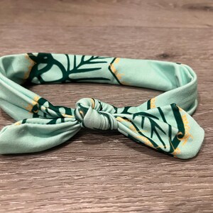 May include: A mint green headband with a knotted bow design. The fabric features a repeating pattern of dark green leaves and yellow accents. The headband is designed to be worn around the head.