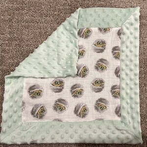 May include: A soft, green baby blanket with a white muslin center featuring a repeating pattern of a cartoon character. The blanket has a soft, textured border.