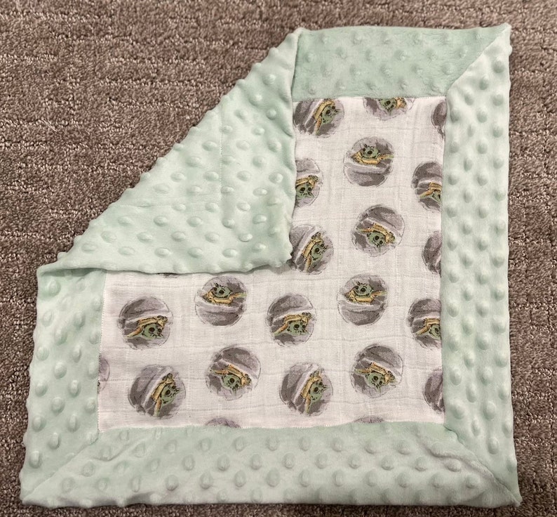 May include: A soft, green baby blanket with a white muslin center featuring a repeating pattern of a cartoon character. The blanket has a soft, textured border.
