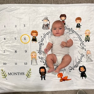 Puede incluir: Una manta de bebé blanca con una tabla de hitos mensuales que incluye los números del 1 al 12. La manta tiene un anillo dorado alrededor del número 6. La manta también presenta un círculo de personajes de El Señor de los Anillos, incluidos Gandalf, Frodo y Gollum. Un bebé está acostado en la manta.