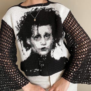 Könnte beinhalten: Ein schwarz-weißer Häkelpullover mit einem Porträt. Der Pullover hat einen Rundhalsausschnitt und lange Ärmel mit einem Netzmuster. Das Porträt zeigt eine Person mit dunklem Haar und heller Haut. Ein einzigartiges Stück tragbarer Kunst.