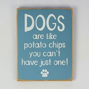 May include: A blue wooden sign with white text that reads "Dogs are like potato chips you can't have just one!" with a white paw print at the bottom.