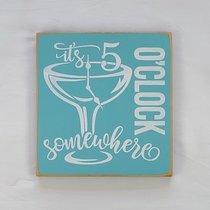 May include: A light blue wooden sign with the text "it's 5 o'clock somewhere" in white. The sign features a white silhouette of a martini glass with clock hands set at 5 o'clock.