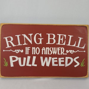 May include: A red wooden sign with white text that reads "Ring Bell If No Answer... Pull Weeds". The sign has a decorative border and green grass accents.