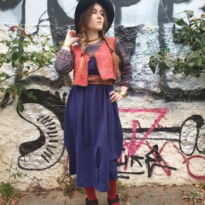 May include: A woman wearing a blue dress, a red vest, and a black hat. She is standing in front of a graffiti wall.