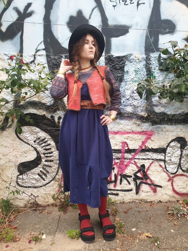 May include: A woman wearing a blue dress, a red vest, and a black hat. She is standing in front of a graffiti wall.