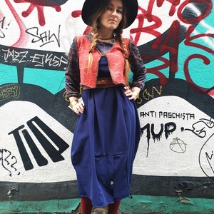 May include: A woman wearing a red vest over a blue dress, a black hat, and red socks. She is standing in front of a graffiti wall with the words "Anti Fascista" and "MUP" written on it.