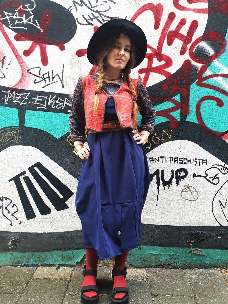 May include: A woman wearing a red vest over a blue dress, a black hat, and red socks. She is standing in front of a graffiti wall with the words "Anti Fascista" and "MUP" written on it.