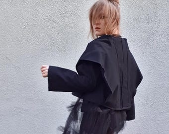 May include: A black jacket with wide sleeves and a cape-like collar. The jacket is paired with a black tulle skirt. The outfit is set against a plain, light-coloured wall, creating a contrast with the dark clothing.