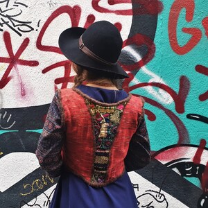 May include: A woman wearing a red, blue, and gold embroidered vest over a blue dress. She is wearing a black hat and standing in front of a graffiti wall.