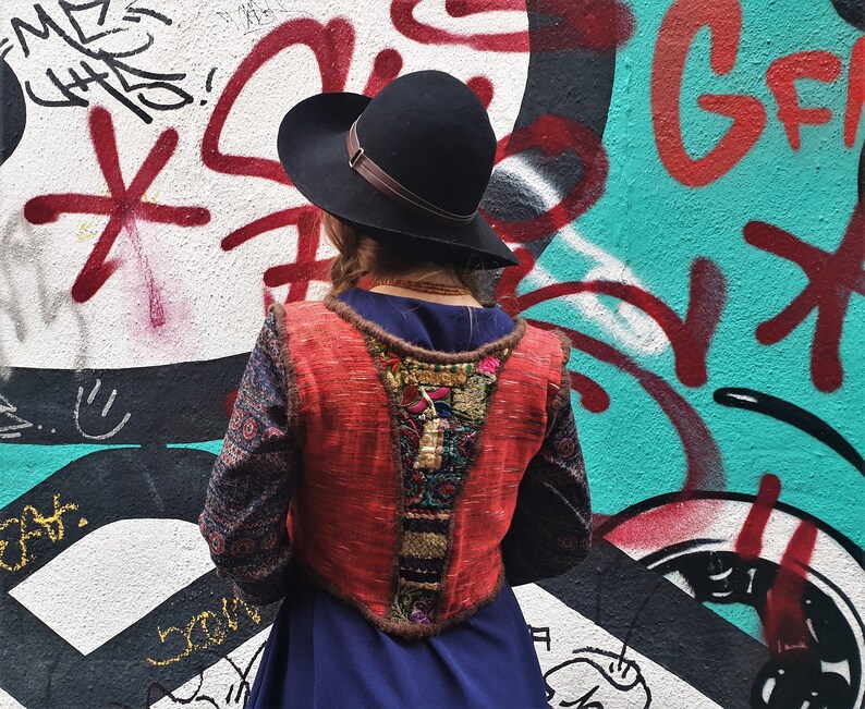 May include: A woman wearing a red, blue, and gold embroidered vest over a blue dress. She is wearing a black hat and standing in front of a graffiti wall.