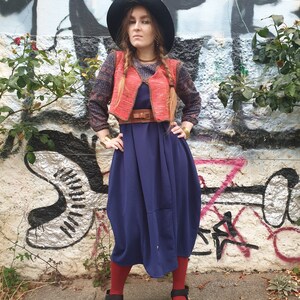 May include: A woman wearing a blue dress, a red vest, a black hat, and red socks. She is standing in front of a graffiti wall.