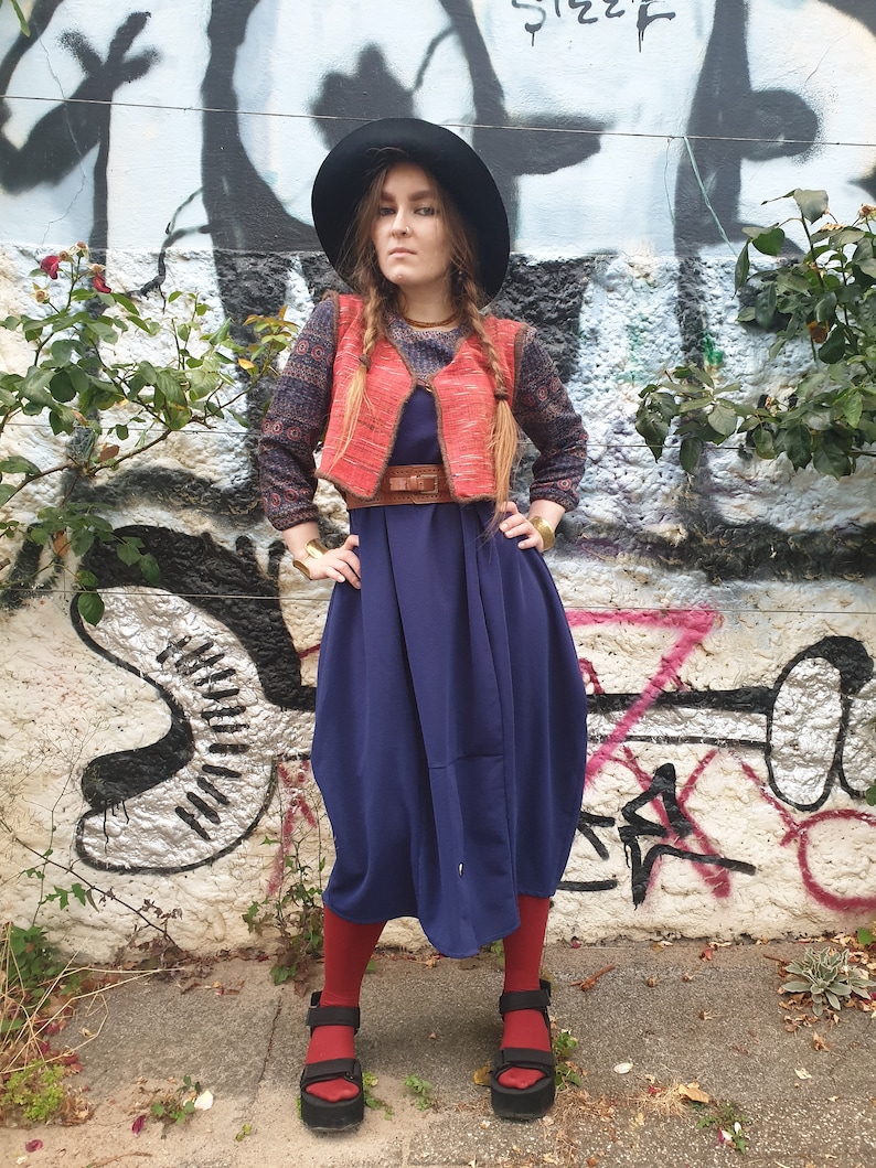 May include: A woman wearing a blue dress, a red vest, a black hat, and red socks. She is standing in front of a graffiti wall.