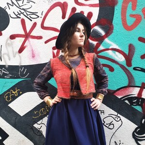 May include: A woman wearing a red vest over a blue patterned long-sleeved shirt, a dark blue skirt, and a black hat. She is standing in front of a graffiti wall.