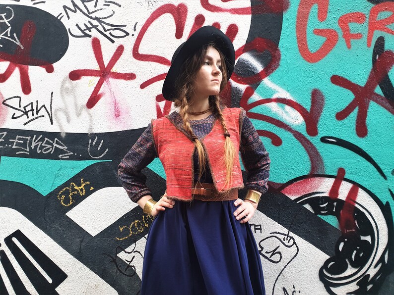 May include: A woman wearing a red vest over a blue patterned long-sleeved shirt, a dark blue skirt, and a black hat. She is standing in front of a graffiti wall.