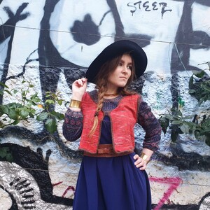 May include: A woman wearing a red and brown knitted vest with a blue and black patterned long-sleeved shirt underneath. She is wearing a wide black hat, a brown belt, and gold bracelets. She is standing in front of a graffiti wall with green plants.