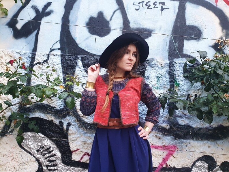 May include: A woman wearing a red and brown knitted vest with a blue and black patterned long-sleeved shirt underneath. She is wearing a wide black hat, a brown belt, and gold bracelets. She is standing in front of a graffiti wall with green plants.