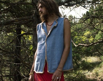 May include: A light blue sleeveless vest with a brown trim and a red and yellow patterned skirt. The vest has a collar and a button closure. The person is standing in a forest.