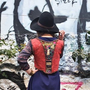 May include: A woman wearing a red and brown embroidered vest with a black hat and a blue dress. The vest has a variety of colorful embroidery and tassels.