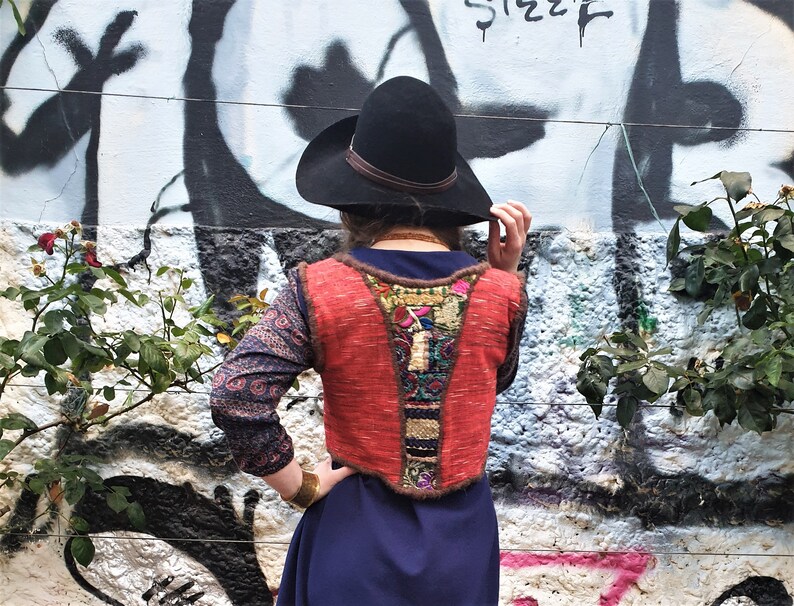 May include: A woman wearing a red and brown embroidered vest with a black hat and a blue dress. The vest has a variety of colorful embroidery and tassels.