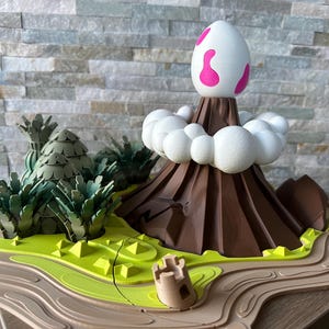 Könnte beinhalten: Ein 3D-gedrucktes Modell einer Vulkaninsel mit einem braunen Berg, einer weißen Wolke und einem rosa Ei auf der Spitze. Die Insel ist von grünen Bäumen und einem braunen Fluss umgeben.
