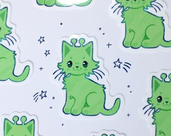 May include: A collection of die-cut stickers featuring a light green cat with antennae, large dark eyes, and a small smile. The cat is surrounded by small dark blue line-art stars and plus signs, creating a repeating pattern of alien-like felines.