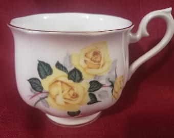 May include: A white teacup with a gold rim and a floral design featuring yellow roses and green leaves.