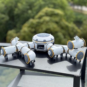 Op de afbeelding: Een wit en grijs ruimtestationmodel met gele accenten. Het model heeft een centrale cirkelvormige hub en verbindende cilindrische modules. De modules hebben kleine zwarte poten en het ontwerp suggereert een futuristisch of sciencefiction thema.