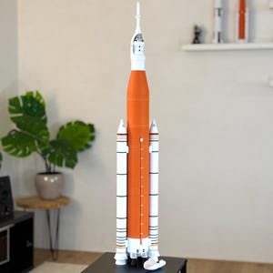 Könnte beinhalten: Ein detailliertes Modell einer Rakete, hauptsächlich orange und weiß, steht auf einer schwarzen Oberfläche. Die Rakete hat mehrere Stufen, Seitenbooster und eine spitze Spitze. Das Modell stellt ein Raumfahrzeug dar.